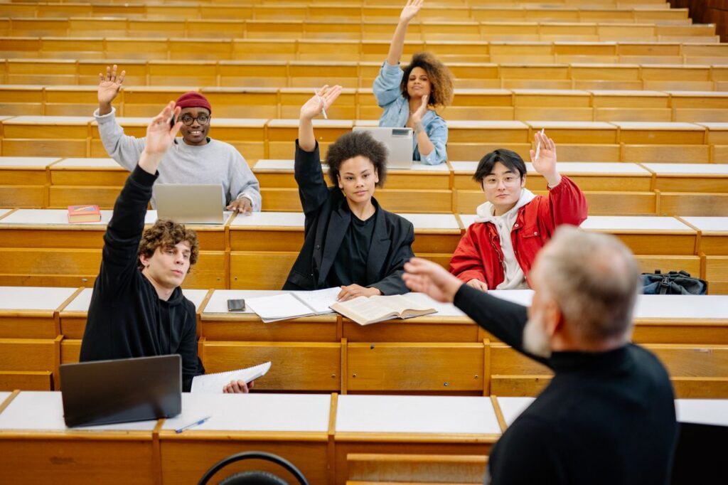 A diverse group of university students participating in a lecture, raising hands to engage with the professor.